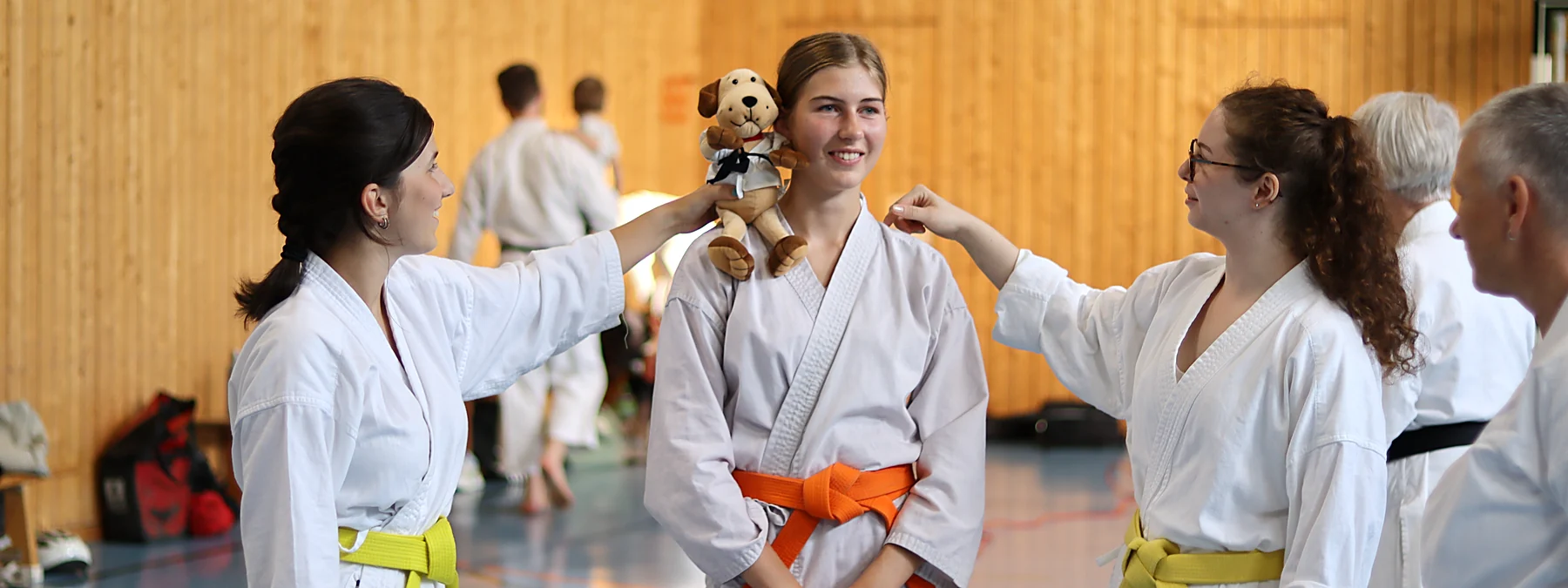 Drei Karateka lachen miteinander, eine setzt der mittleren einen Stoffhund im Karate-Anzug auf die Schulter