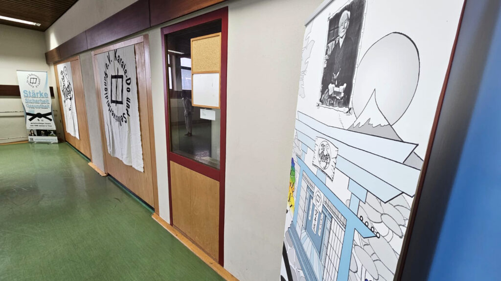 An einem Ende der Halle sind Aufsteller und Stoffbahnen zu sehen mit einem Bild von Funakoshi, dem Vereinssymbol, einem Shotokan-Tiger und der Schrift "Stärke, Sicherheit, Gesundheit".